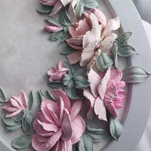 May include: A decorative, oval-shaped wall art piece featuring a relief design of pastel-colored flowers and leaves. The flowers are primarily pink and white, with green leaves, set against a gray background.