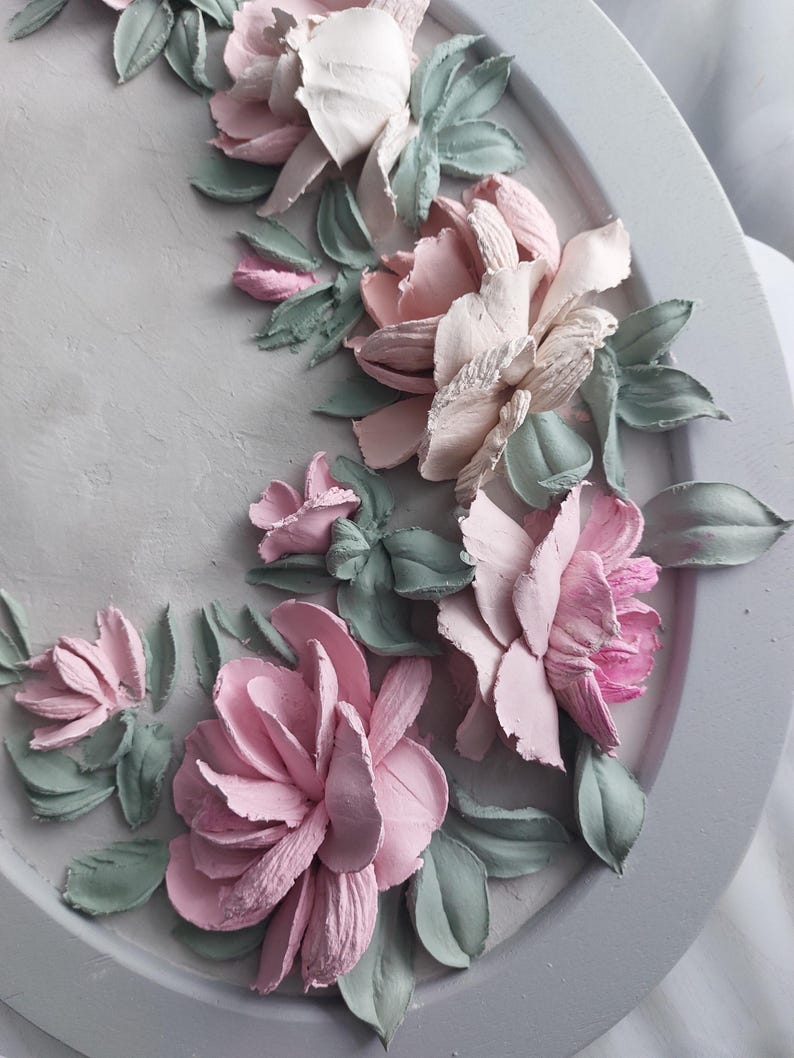 May include: A decorative, oval-shaped wall art piece featuring a relief design of pastel-colored flowers and leaves. The flowers are primarily pink and white, with green leaves, set against a gray background.