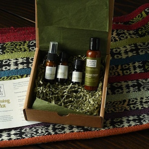 May include: A brown cardboard box filled with green and white shredded paper. Inside the box are four small bottles of skincare products. The bottles are labelled "Clear Deep", "Tea Tree", "Rose & Lavender", and "Algae Deep". The box is labelled "Source Vital Apothecary Acne Cleansing Starter Kit".