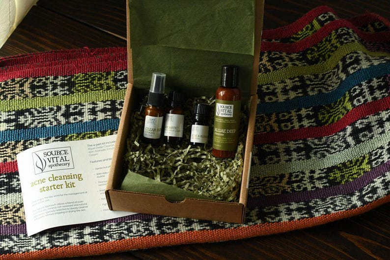 May include: A brown cardboard box filled with green and white shredded paper. Inside the box are four small bottles of skincare products. The bottles are labelled "Clear Deep", "Tea Tree", "Rose & Lavender", and "Algae Deep". The box is labelled "Source Vital Apothecary Acne Cleansing Starter Kit".