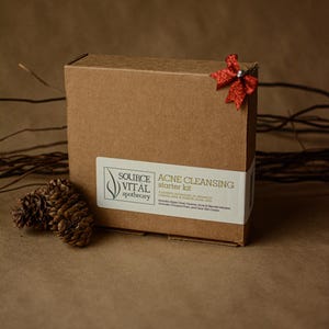 May include: A brown cardboard box with a red bow on top. The box is labelled "ACNE CLEANSING starter kit" and features the logo "SOURCE VITAL apothecary".