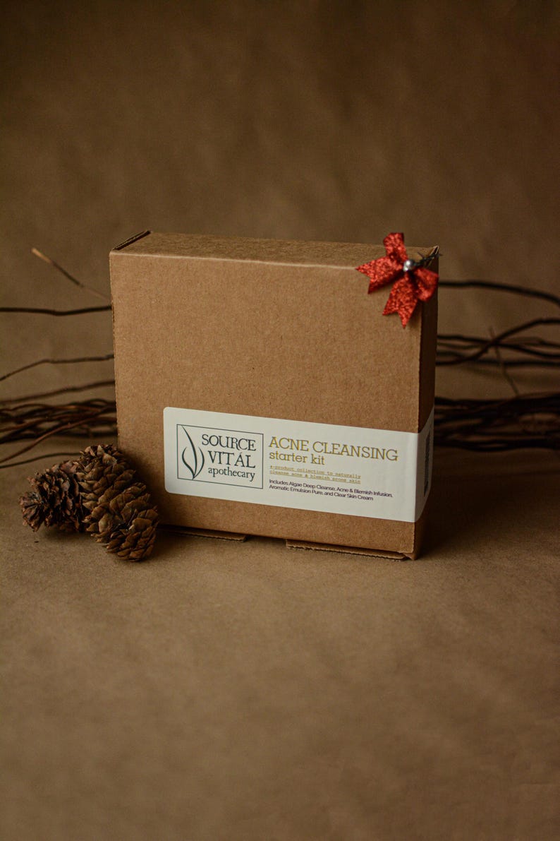 May include: A brown cardboard box with a red bow on top. The box is labelled "ACNE CLEANSING starter kit" and features the logo "SOURCE VITAL apothecary".