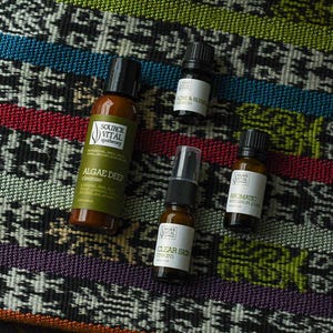 May include: Four small bottles of essential oils and skincare products on a colourful patterned fabric. The bottles are labelled "Algae Deep Conditioner", "Clear Skin Serum", "Aromatic Blend", and "Acne & Balance".