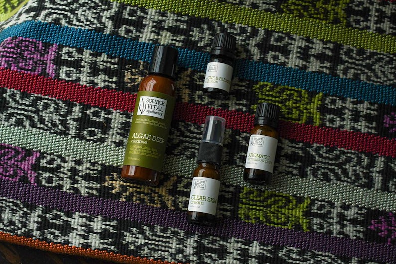 May include: Four small bottles of essential oils and skincare products on a colourful patterned fabric. The bottles are labelled "Algae Deep Conditioner", "Clear Skin Serum", "Aromatic Blend", and "Acne & Balance".