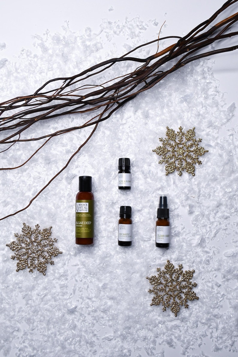 May include: Four small bottles of essential oils and a lotion bottle on a white surface with gold snowflake decorations. The bottles are labelled "Clear Skin", "Aromatic", "Algae Deep", and "Rose & Frankincense".