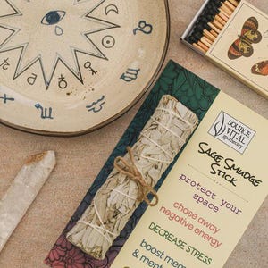 Puede incluir: Un surtido de artículos espirituales, que incluyen un plato de cerámica beige con símbolos del zodíaco, un palo de salvia, un cristal de cuarzo transparente y una caja de cerillas con ilustraciones de mariposas. El palo de salvia va acompañado de una tarjeta con texto.