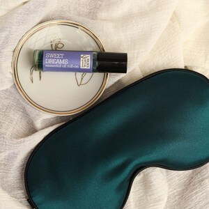 May include: A green silk sleep mask with a black trim, a small white saucer with a gold rim, and a glass bottle of essential oil roll-on labelled "Sweet Dreams" on a white fabric background.