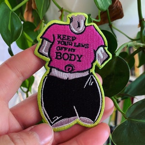 May include: A pink and black embroidered patch with the text "Keep your laws off my body". The patch is shaped like a person wearing a t-shirt and shorts.