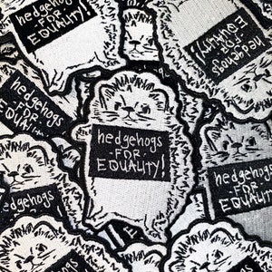 Pode incluir: Uma coleção de patches bordados em branco e preto com um design de ouriço. Cada patch tem o texto "hedgehogs FOR EQUALITY!" em negrito. Os patches estão densamente embalados.