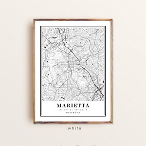 May include: A framed black and white map print of Marietta, Georgia. The map features intricate road and street details, with the city name and coordinates displayed at the bottom. The frame is a light brown color, and the word "white" is printed at the bottom.