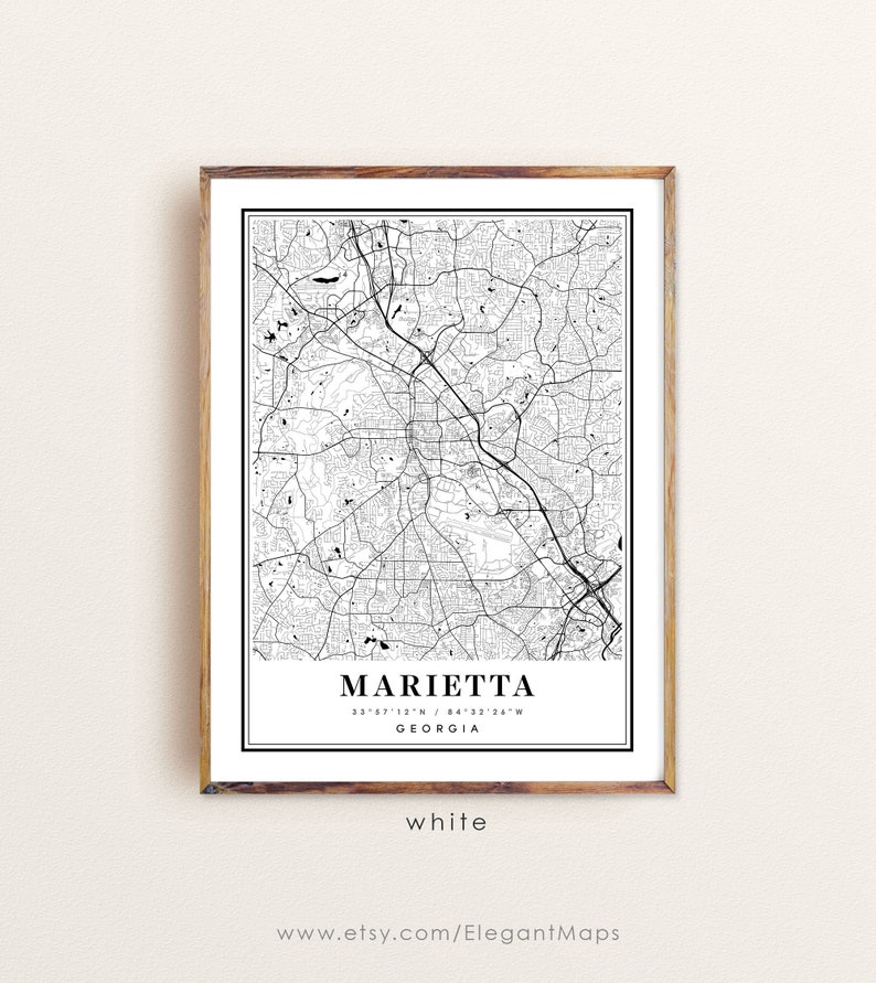 May include: A framed black and white map print of Marietta, Georgia. The map features intricate road and street details, with the city name and coordinates displayed at the bottom. The frame is a light brown color, and the word "white" is printed at the bottom.