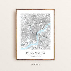 May include: A black and white map print of Philadelphia, Pennsylvania with the city's name and coordinates in a minimalist font. The map is framed in a light brown wood frame. The text "modern" is printed below the map and a website address is printed below the text.
