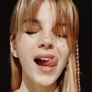 May include: A young woman with long blonde hair and bangs, wearing a yellow tank top, sticks her tongue out and smiles. She has glitter on her face and is wearing small earrings.