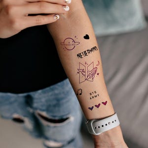 May include: A person's arm with several temporary tattoos. The tattoos include a black heart, the words "NEVERMIND", a stylized "B" with a whale inside, the words "BTS ARMY", and three pink hearts.