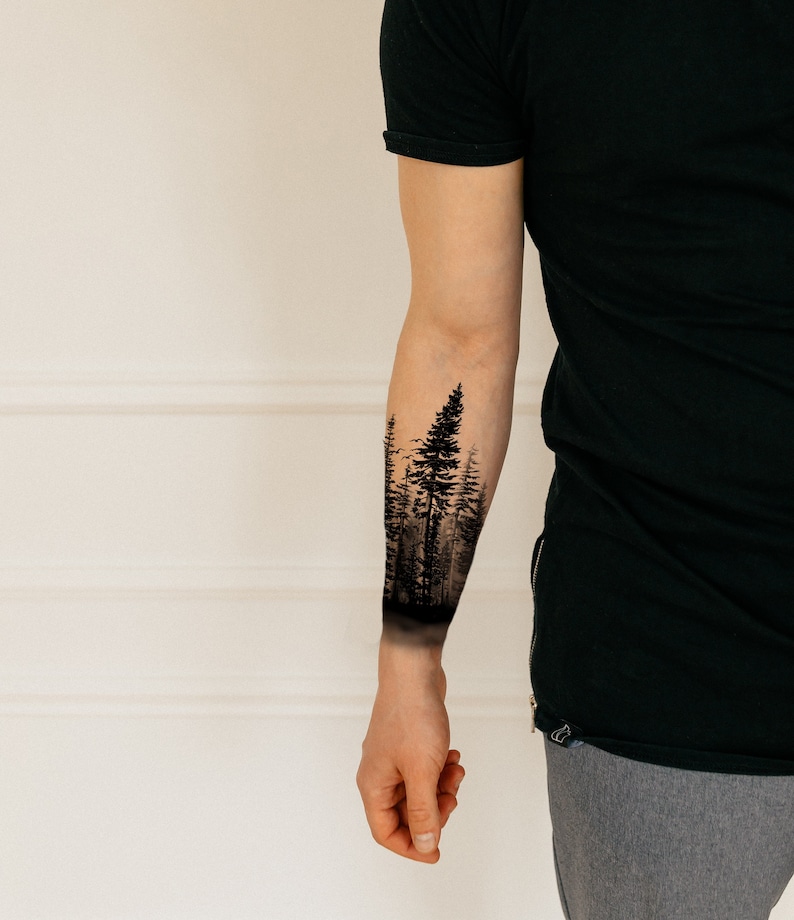 Op de afbeelding: Zwart-wit bos tattoo ontwerp op een arm. De tattoo toont een gedetailleerde weergave van hoge dennenbomen, waardoor een silhouet ontstaat tegen een mistige achtergrond. Het ontwerp bedekt de onderarm en toont een op de natuur ge&iuml;nspireerd thema.