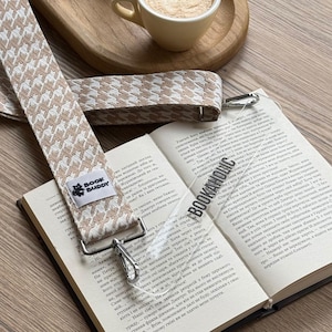 May include: Open book with a houndstooth patterned book strap and a clear bookmark that reads "BOOKAHOLIC". A cup of coffee sits on a wooden tray in the background. The book strap has a "BOOK BUDDY" label.