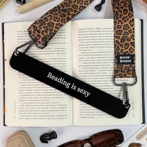 Puede incluir: Marcapáginas negro con el texto "Reading is sexy" en blanco. El marcapáginas tiene una correa con estampado de leopardo y un cierre plateado, colocado sobre las páginas de un libro.