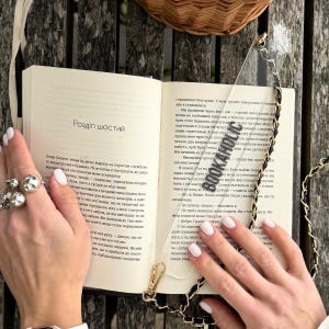 以下が含まれることがあります： 「BOOKAHOLIC」の文字と黒いチェーンストラップが付いた透明なアクリルブックマークが、開いた本の中に置かれています。本はウクライナ語のテキストが書かれたページが開かれています。シーンは木製の表面に設定されています。