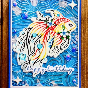 May include: A handmade birthday card featuring a colorful fish design. The fish has red, yellow, and white details with black outlines, adorned with blue flowers. The card includes the text "happy birthday" and is embellished with blue gems and white star shapes.