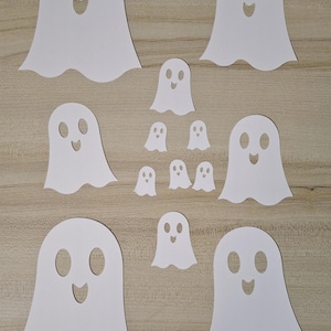 May include: A collection of white paper ghost cutouts in various sizes, arranged on a light brown wooden surface. Each ghost has a simple, smiling face with oval eyes and a curved mouth, perfect for Halloween decorations.