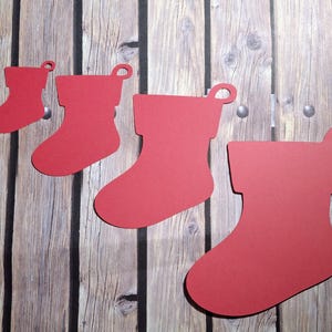 May include: Red Christmas stocking cutouts in various sizes, arranged on a wooden background. Each stocking has a loop at the top. The stockings are made of a solid red material and are in different sizes.