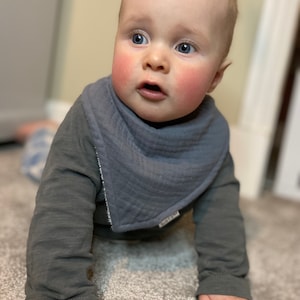 May include: A baby wearing a gray long-sleeved shirt and a gray bib. The bib is a bandana style with a white stitched edge. The baby has fair skin, blue eyes, and reddish-blond hair.