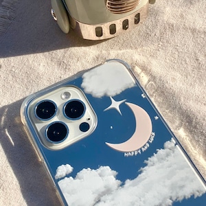 Puede incluir: Una funda transparente para teléfono con un cielo azul, nubes blancas, una luna creciente rosa y una estrella blanca. El texto "Happy and Luck" está impreso en la funda.