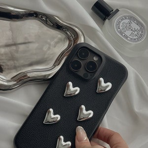May include: Black phone case with silver heart-shaped embellishments. The case is made of a textured material and has a camera lens cutout.