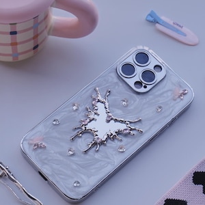 Puede incluir: Funda de teléfono plateada con diseño de mariposa, gemas transparentes y acabado nacarado. La funda está junto a una taza rosa, un dije en forma de melocotón y una pinza para el pelo. También se ve un calcetín rosa y negro.