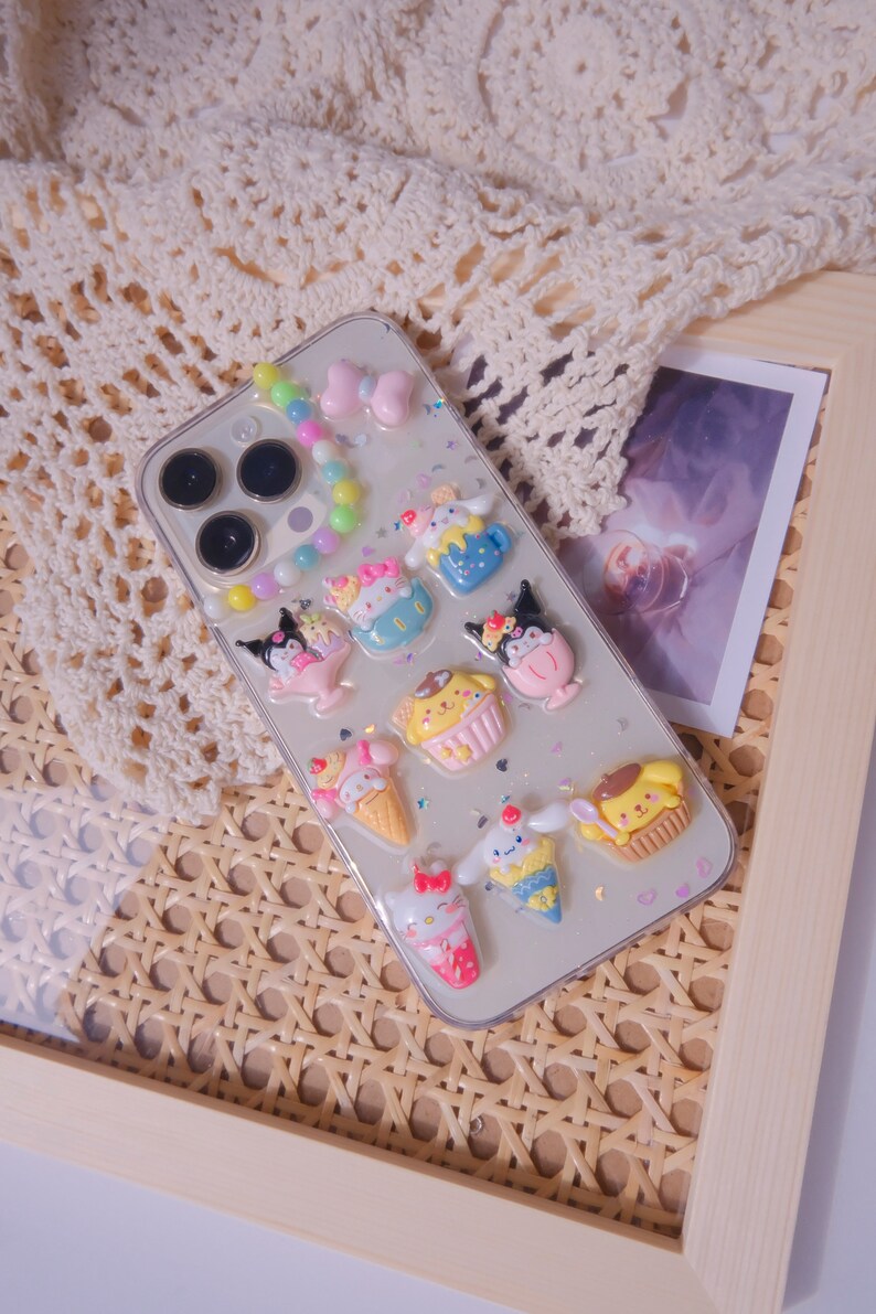 May include: A clear phone case with a white background and colorful cartoon characters. The characters are holding ice cream cones, cupcakes, and other treats. There are also colorful beads and glitter on the case.