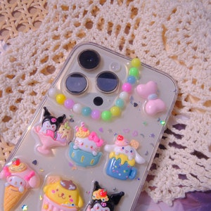 May include: A clear phone case with a colorful beaded charm and various cartoon character charms. The charms are of different colors and shapes, including a pink cupcake, a blue ice cream cone, and a yellow ice cream cup. The case is decorated with glitter and stars.