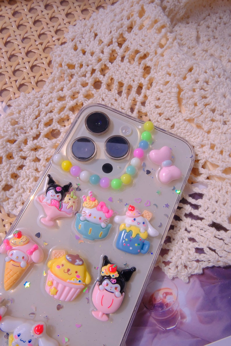 May include: A clear phone case with a colorful beaded charm and various cartoon character charms. The charms are of different colors and shapes, including a pink cupcake, a blue ice cream cone, and a yellow ice cream cup. The case is decorated with glitter and stars.