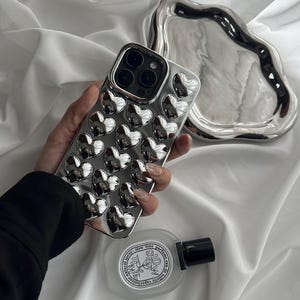 Puede incluir: Una funda de teléfono plateada con un diseño de corazón. La funda está hecha de un material brillante y reflectante y tiene un patrón de corazón en relieve. La funda está siendo sostenida en una mano.