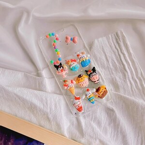 May include: A clear phone case with colorful charms attached. The charms include a bow, a cupcake, an ice cream cone, and a cartoon character.