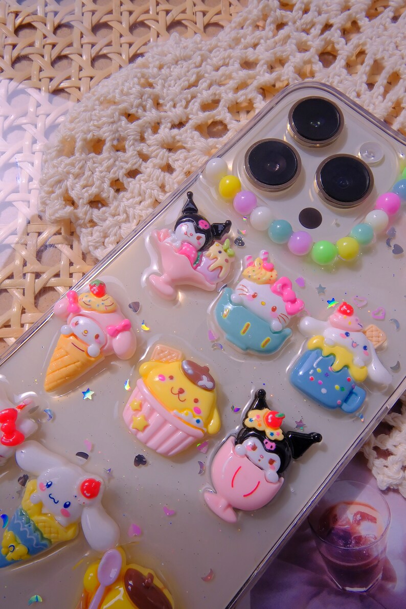 May include: A clear phone case with a variety of colorful cartoon characters and a beaded charm. The characters include a cat, a dog, a rabbit, and a bear. The case is decorated with glitter and stars.