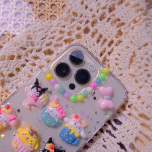 May include: A clear phone case with a colorful beaded charm and various cartoon character charms. The charms are in the shape of a cupcake, a cat, a dog, and a bear. The phone case is decorated with glitter and hearts.