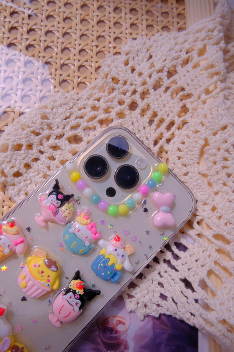 May include: A clear phone case with a colorful beaded charm and various cartoon character charms. The charms are in the shape of a cupcake, a cat, a dog, and a bear. The phone case is decorated with glitter and hearts.
