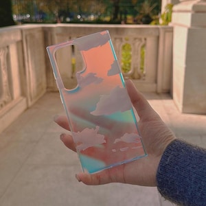 Könnte beinhalten: Eine transparente Handyhülle mit Wolkenmuster. Die Hülle hat eine rechteckige Form mit Aussparungen für Kamera und Knöpfe. Die Wolken sind weiß und der Hintergrund hat einen Farbverlauf aus Rosa, Orange und Blau.