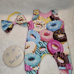 May include: A baby outfit and matching bow featuring a colorful donut pattern. The one-piece outfit has a pink neckline and cuffs, with a variety of donuts in shades of blue, pink, and brown. The bow is on a yellow elastic band. A wooden disc reads "18-19" Tiny Baby".