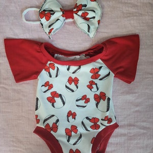 May include: A white baby onesie with a red and white baseball print. The onesie has red short sleeves and a red trim around the neckline. A red and white bow headband is included.