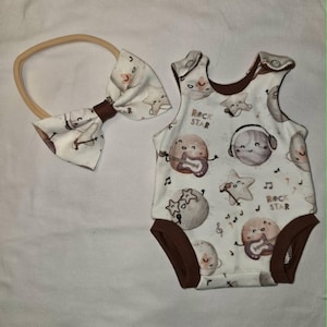 May include: A white baby bodysuit and matching bow headband set. The bodysuit features a print of smiling planets, stars, and musical notes, with the text "ROCK STAR". The trim is brown. The bow is attached to a beige elastic band.