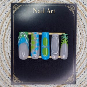 Op de afbeelding: Een set van vijf kunstnagels met verschillende ontwerpen. De nagels hebben een heldere basis met ontwerpen zoals blauwe en groene wervelingen, een blauwe basis met een groene ovaal en een zilveren omtrek. De tekst "Nail Art" staat bovenaan.