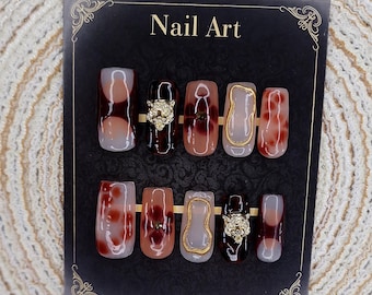 Może przedstawiać: Zestaw sztucznych paznokci ułożonych na czarnej karcie z napisem "Nail Art". Paznokcie mają różne wzory, w tym zwierzęcy nadruk, złote akcenty i złotą głowę lwa. Paznokcie są w odcieniach brązu, beżu i czerwieni.