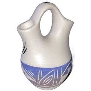May include: A white ceramic wedding vase with a blue and white geometric design around the base. The vase has a handle and two spouts. Black lines are painted on the side of the vase.