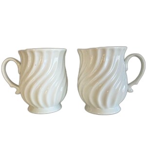 May include: Two off-white ceramic mugs with a swirled design. Each mug has a handle and a slightly flared rim. The mugs are identical in shape and size, and are set against a white background.