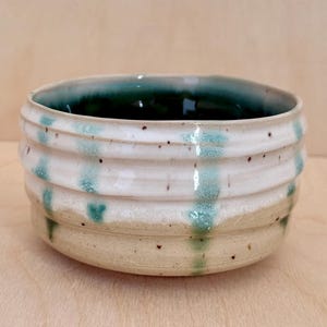 May include: A handmade ceramic bowl with a white exterior and a dark green interior. The bowl features horizontal ridges and turquoise drips. The bottom is a speckled tan color. This unique bowl is perfect for serving food or as a decorative piece.