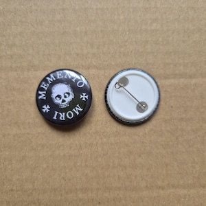 May include: Black and white button with a skull and crossbones and the text "Memento Mori" in a circle. The back of the button is white with a safety pin.