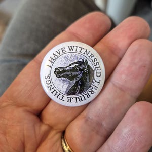 May include: A circular white pin-back button featuring a black and white illustration of a horse head in the centre. The text "I HAVE WITNESSED TERRIBLE THINGS." is printed in black capital letters around the perimeter of the circular button design.