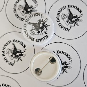 May include: A set of six white round button badges with black text and images. The badges feature a black and white illustration of a person riding a demon. The text on the badges reads "Read Banned Books".