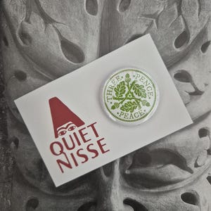 May include: A white card with the text "QUIET NISSE" in red, and a red hat graphic. A round button with green text and a green leaf design that says "THREE PENCE PEACE". The background is a grey stone carving.